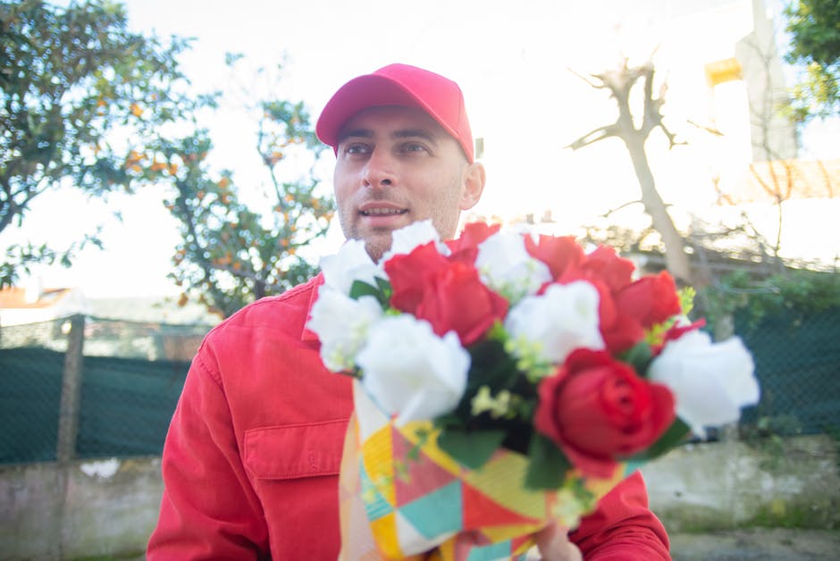 A florist wearing a red cap and red jacket is outdoors, holding a vibrant floral bouquet composed of red and white flowers, including roses and lilies, wrapped in colorful paper. The bouquet is positi