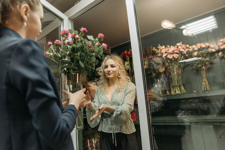A young woman with curly blonde hair, dressed in a light-colored floral blouse, stands inside a flower shop near a glass door, receiving a bouquet of fresh pink roses wrapped in brown kraft paper from