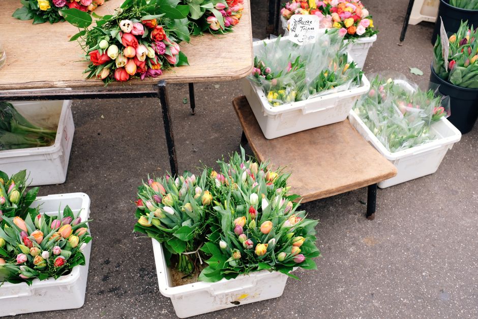 Several bouquets of fresh tulips with pink, white, yellow, and red petals, arranged with green foliage, are displayed in white plastic crates and placed on a wooden table and small wooden platform out