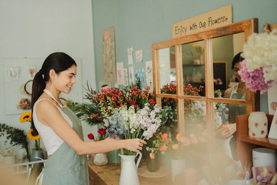 A young woman with dark hair tied back, dressed in a white top and light green apron, arranging a tall white vase filled with a fresh floral bouquet that includes red, white, purple, and pink blossoms