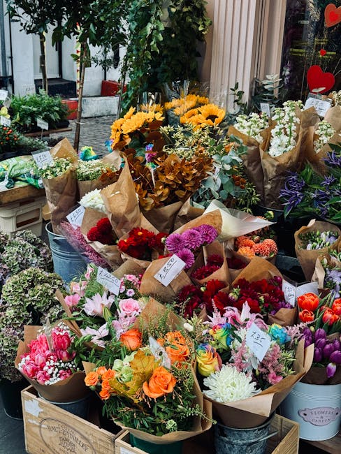 A display of fresh flowers arranged in various bouquets and wrapped in brown kraft paper, featuring a vibrant mix of colors including pink, orange, red, purple, and white blooms. The bouquets are arra