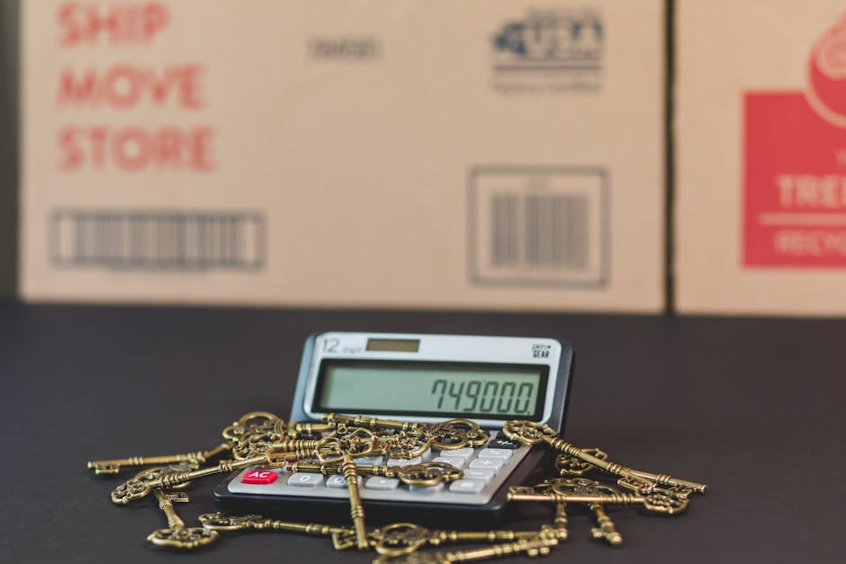 A calculator displaying the number 749800 is placed on a black surface, surrounded by a pile of antique gold-colored keys, with blurred cardboard boxes in the background. The setting suggests a focus