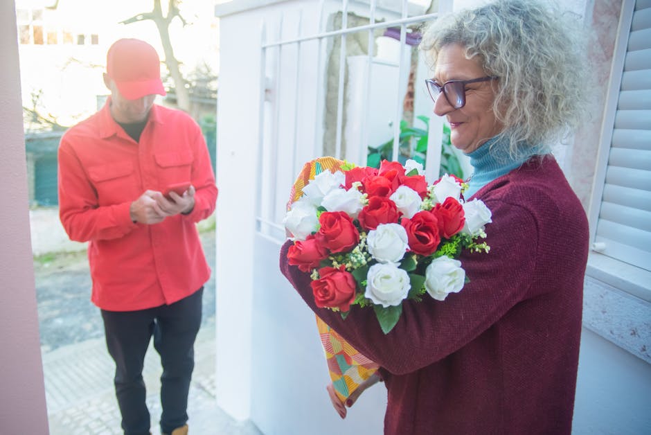 A woman with curly gray hair, wearing glasses, a turquoise turtleneck, and a maroon sweater, is standing in a porch area holding a vibrant floral gift consisting of red and white roses arranged in a r