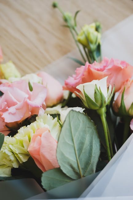A close-up of a floral arrangement featuring soft pink and cream lisianthus flowers with ruffled petals, complemented by fresh green leaves and stems. The bouquet is partially wrapped in light grey pa