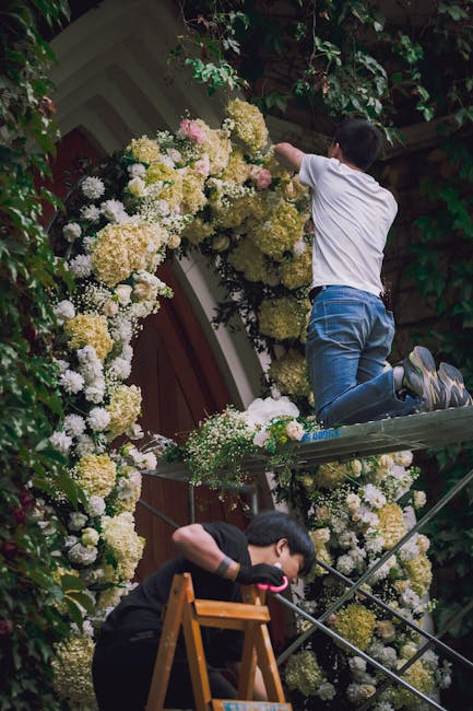 A child in a white t-shirt and blue jeans kneels on a scaffold, adjusting a large, elaborate floral arrangement adorning a building's exterior wall. The arrangement consists of densely packed white an