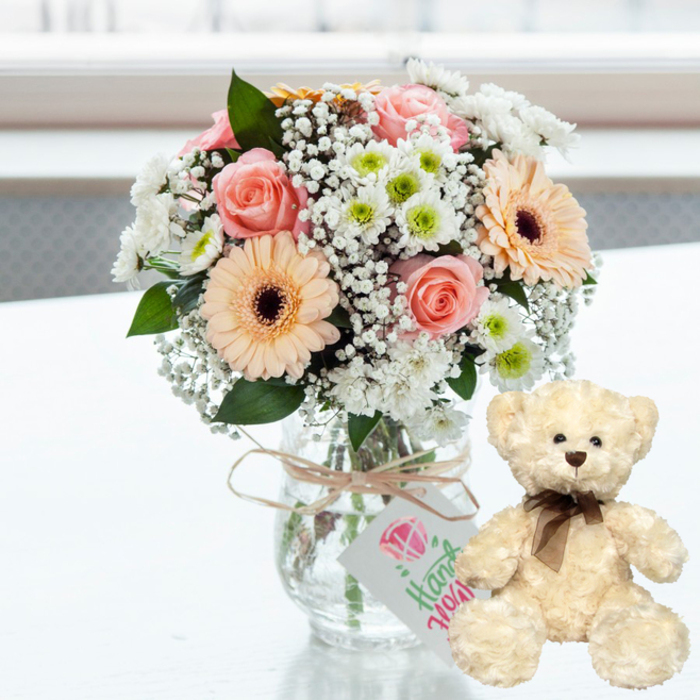 A tender anniversary gift arrangement is captured on a gleaming white surface, the kind of bright, uncluttered backdrop you might find in a modern Harringay apartment near Finsbury Park. At the centre of the image, a generous bouquet spills from a clear glass jar-style vase, its transparency revealing clean stems gathered neatly in water. The palette is composed of soft, romantic pastels: blush pink roses with gently unfurling petals, peach gerbera daisies with expressive dark chocolate centres, and domes of white chrysanthemums edged with a hint of fresh lime-green. Between these larger flowers, airy sprigs of white baby's breath thread and cluster, adding a delicate, cloud-like softness that makes the arrangement feel both full and light. The vase is dressed in a simple, rustic detail-a piece of natural, light brown twine tied snugly around the neck. Suspended from the twine is a small white rectangular tag, decorated with a pink jewel-shaped icon and graceful green handwriting that reads "Handmade," hinting at a personal, crafted-with-love touch from a local florist. To the right of the bouquet, a plush cream teddy bear sits upright, legs stretched slightly forward, as if patiently waiting to be hugged. Its fur appears soft and a little fluffy, with dark brown eyes, a button nose, and a neatly tied brown satin ribbon at the neck that brings a subtle sense of occasion. The background is softly out of focus, suggesting daylight streaming in from a window and bathing the scene in a clean, luminous glow. Together, flowers and bear create a warm, comforting impression-perfect for an anniversary surprise sent across Harringay, whether to rekindle romance, celebrate shared years, or offer a gentle reminder of affection.